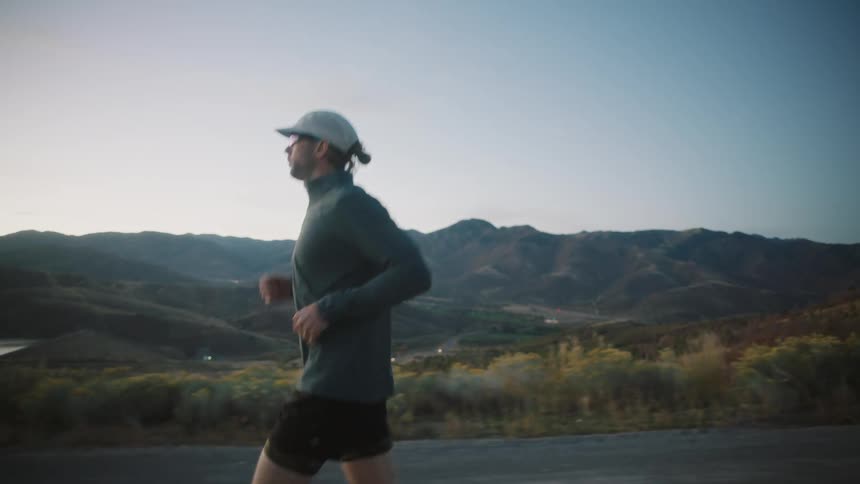 A man wearing a gray jacket black shorts and a gray cap running along a road with a mountain range and a dusky sky in the background