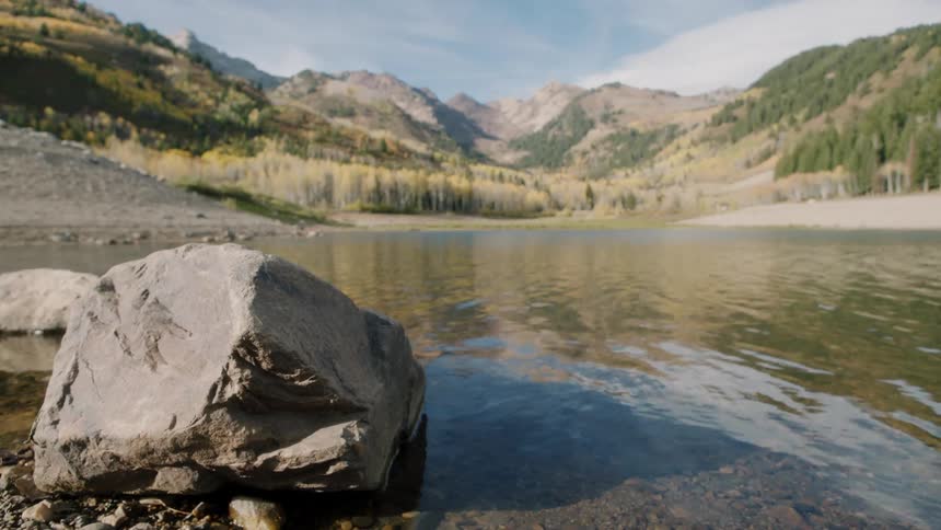 A wide shot captures a large rock at the edge of a clear lake surrounded by mountains and autumn trees under a blue sky