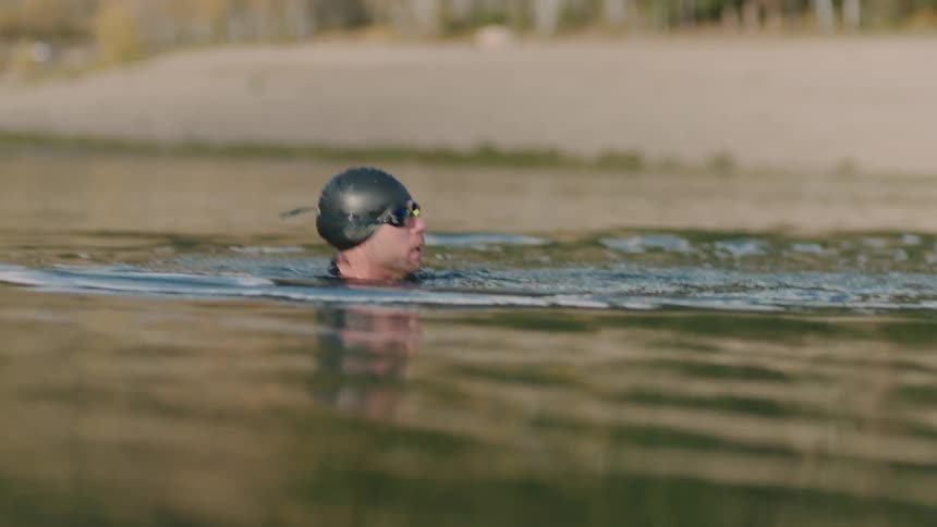 A person wearing a swim cap is swimming in a calm lake with a sandy shore and trees in the background