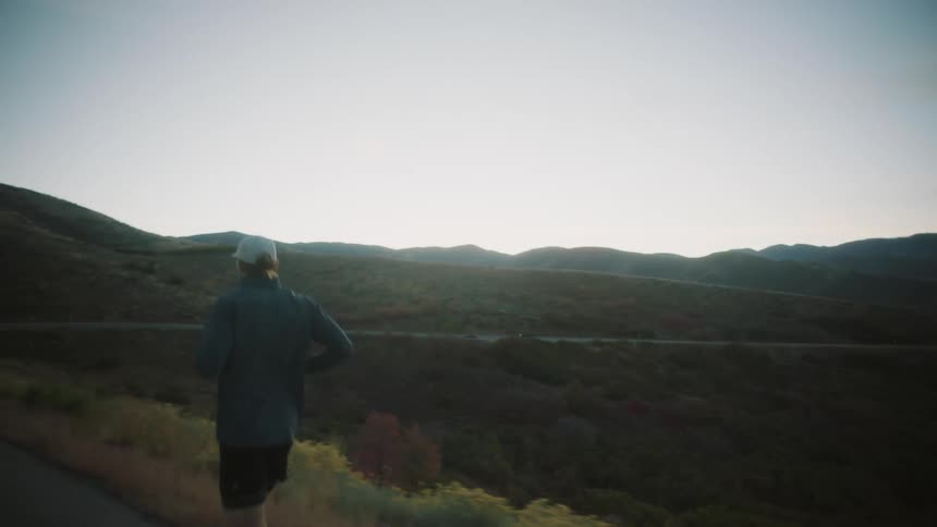 A man is running along a road in a hilly rural landscape during early morning or late evening with the sun low in the sky