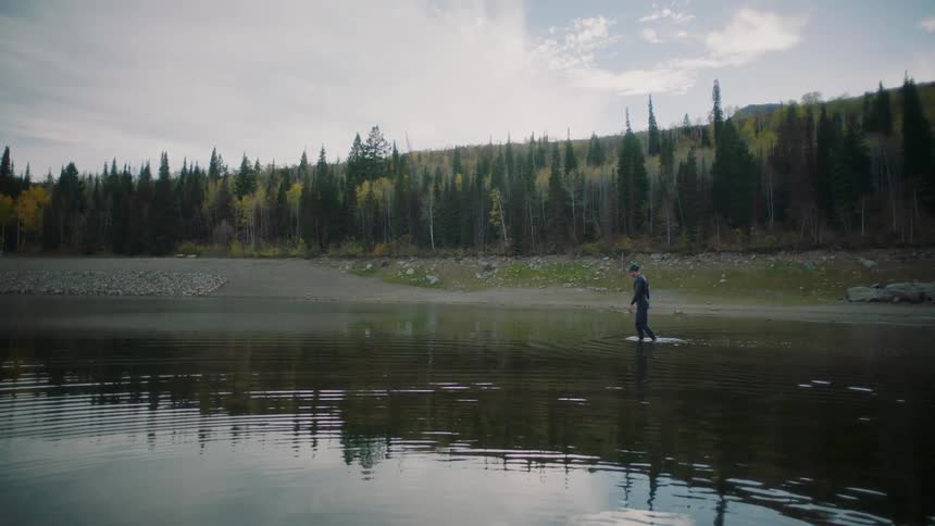 A man wearing a wetsuit wades through a lake on a mostly cloudy autumn day