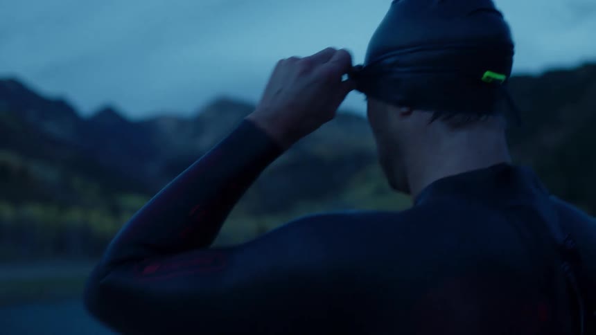 Man swimming in a wetsuit faces the mountainous horizon at dawn facing a lake