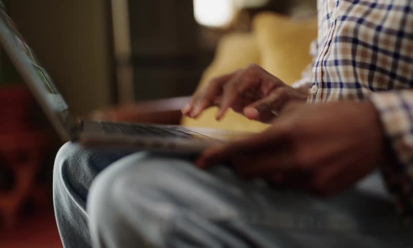 A person in a checkered shirt is sitting on the sofa and opening the laptop computer and then typing