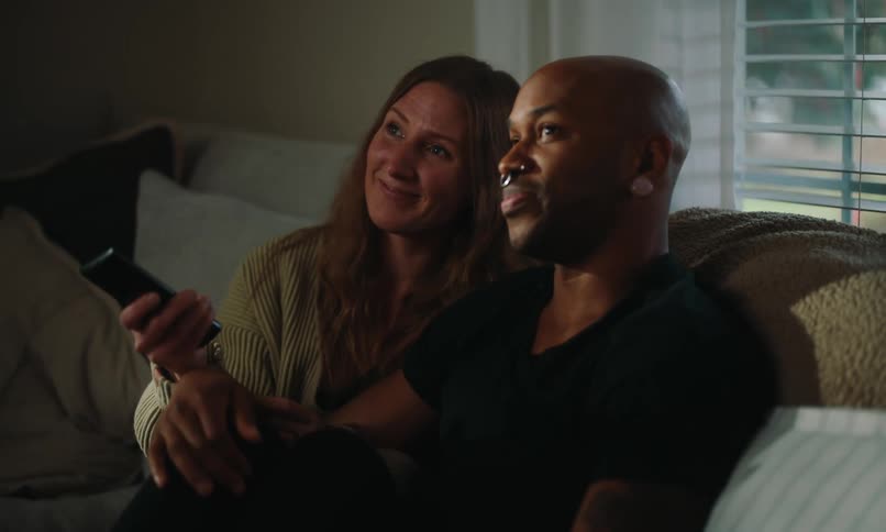 Two people are sitting on a couch one holding a remote control with their hands touching