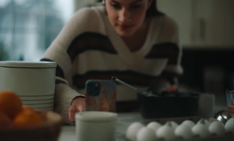 A woman wearing a striped knit sweater is preparing breakfast in a kitchen while looking at a recipe on her smartphone