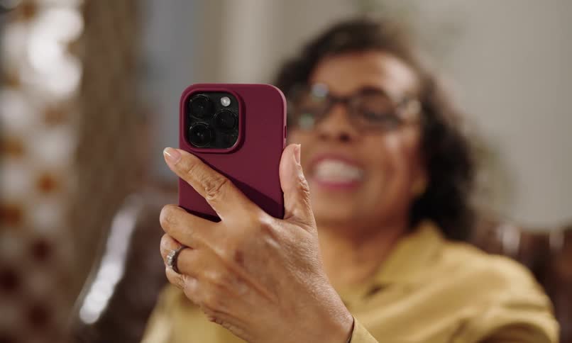A woman with glasses and a brown shirt is on a video call on her iphone which has a red case