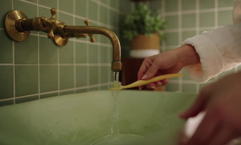 A person in a white robe holds a yellow toothbrush under a brass faucet in a green tiled bathroom