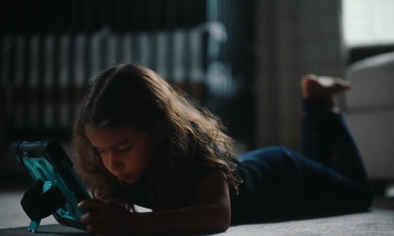 A little girl lying on the floor is looking at a tablet that is in front of her