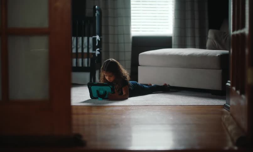 Girl with wavy hair lies on the rug on the wooden floor and uses a tablet in the living room