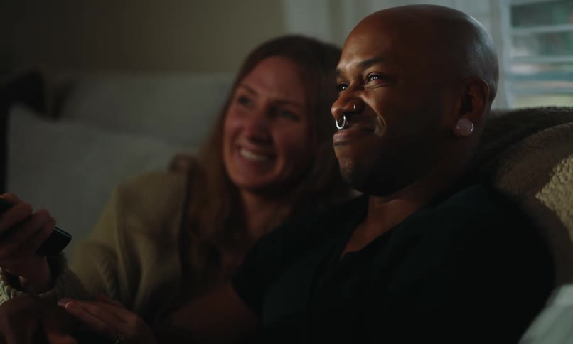 A couple sits together on a couch at home at night changing channels on the television with a remote control