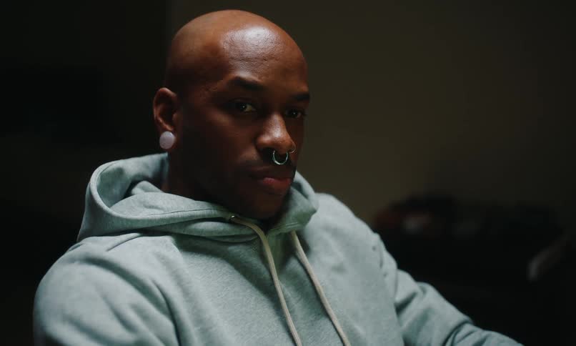 A bald man with a nose ring and a large earring in his lobe wearing a blue hoodie sits in a dimly lit room