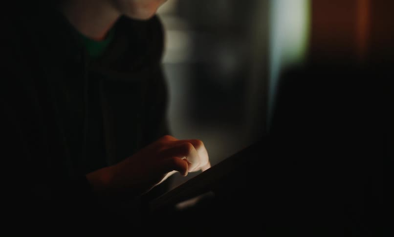 A person is using a tablet in a dimly lit room with their face illuminated by the screen