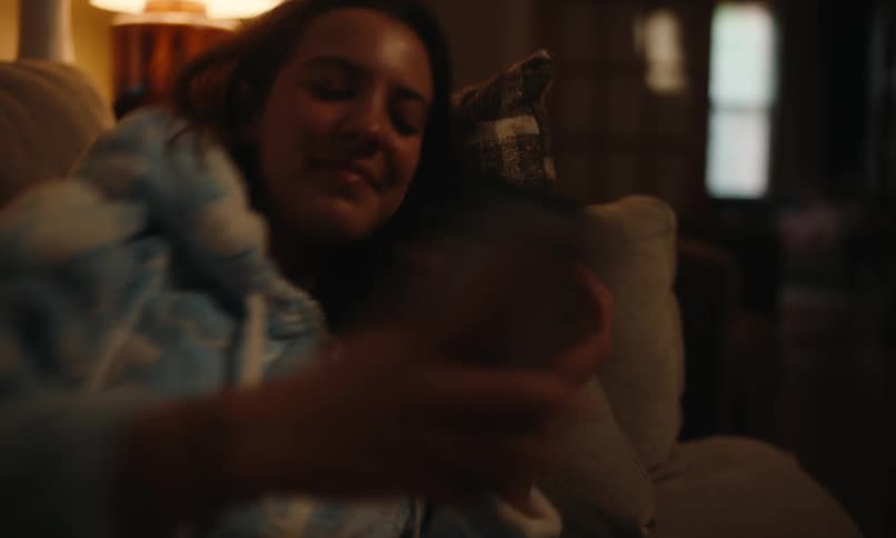 A young woman is sitting on a couch in dim lighting smiling while looking at her phone dressed in a cozy hoodie with a patterned pillow behind her