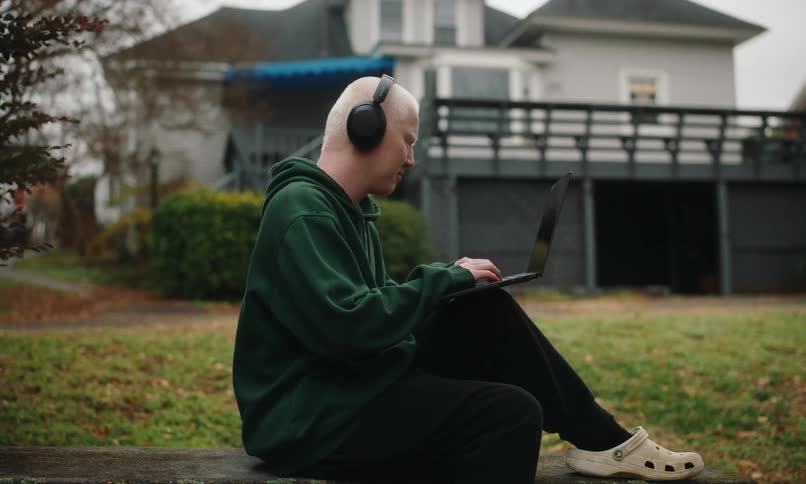 A man with short platinum blonde hair wearing headphones and a dark green hoodie sitting on the grass with a laptop in their lap focused on their work with a suburban house in the background