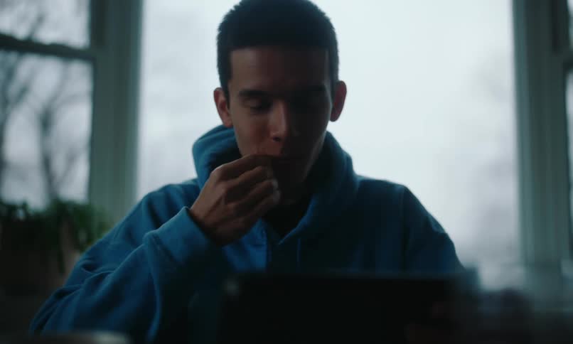 A boy wearing a blue sweater smiles as he eats and watches a tablet
