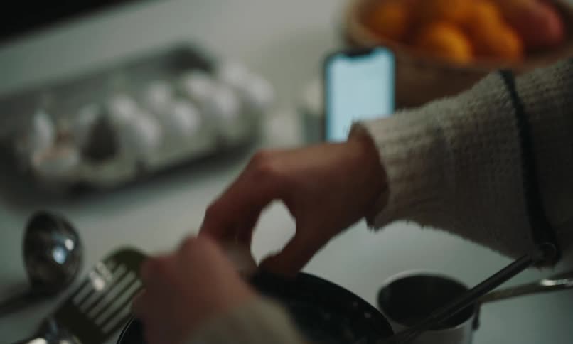 She cracks eggs into a bowl and mixes them with a phone displaying a recipe resting nearby and a carton of broken eggs and a bowl of oranges in the background