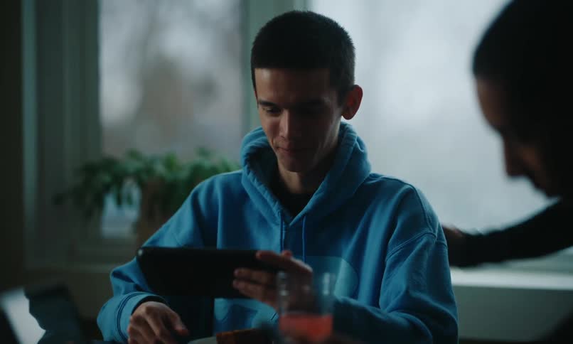 A teenager in a blue hoodie holds a tablet while having breakfast with family or friends