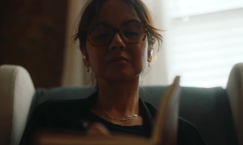 A woman wearing glasses is writing in a notebook seated in a softly lit room with a calm and focused demeanor