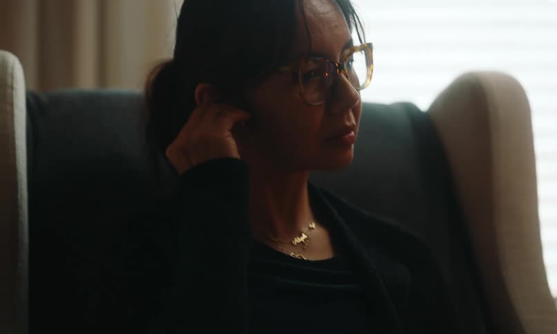 A woman picks up a case with earbuds opens it and puts them in her ears while sitting on a chair in a dimly lit room