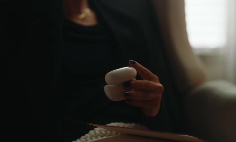 A woman sits in a comfortable chair at home with a blanket and a book on her lap as she puts on earbuds