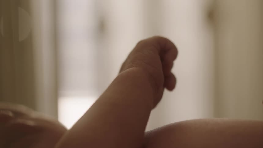 A baby lying on its back its arm raised with a clenched fist against a neutral toned background