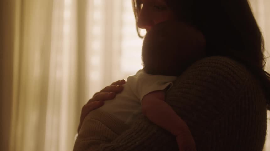 A mother is holding a baby in a warmly lit room with curtains in the background