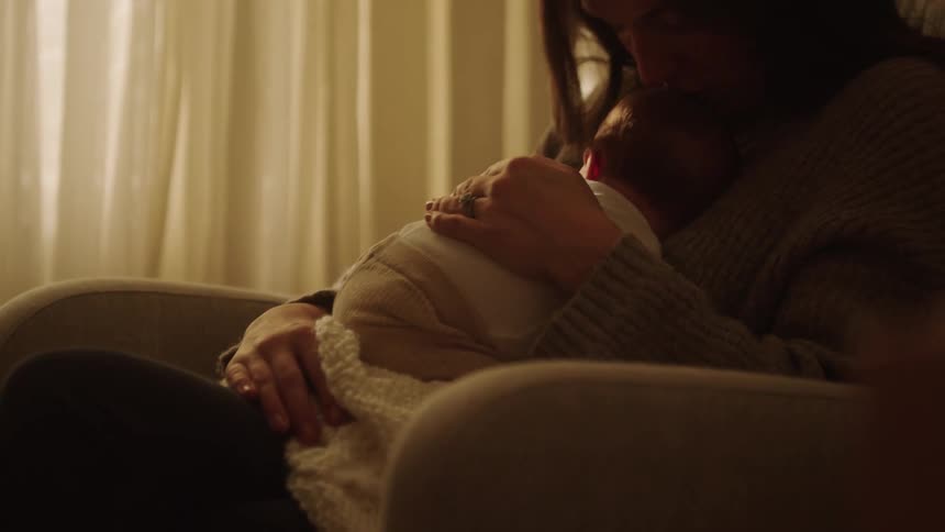 A warm intimate moment between a mother and her baby in a softly lit room where she cradles the infant close to her chest wrapped in a cozy blanket conveying deep affection and tenderness