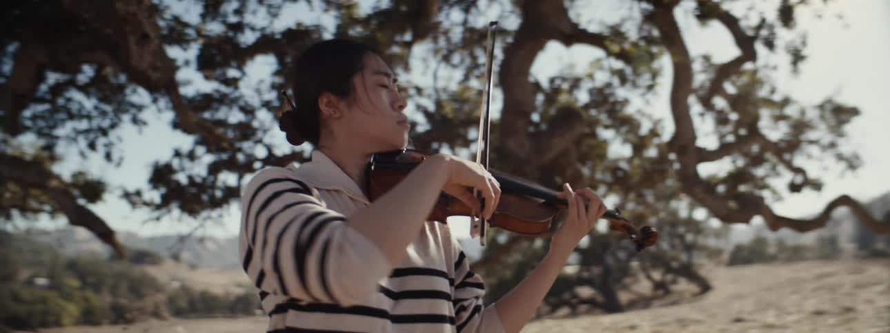 A man with his eyes closed playing the violin next to a large tree on a sunny day