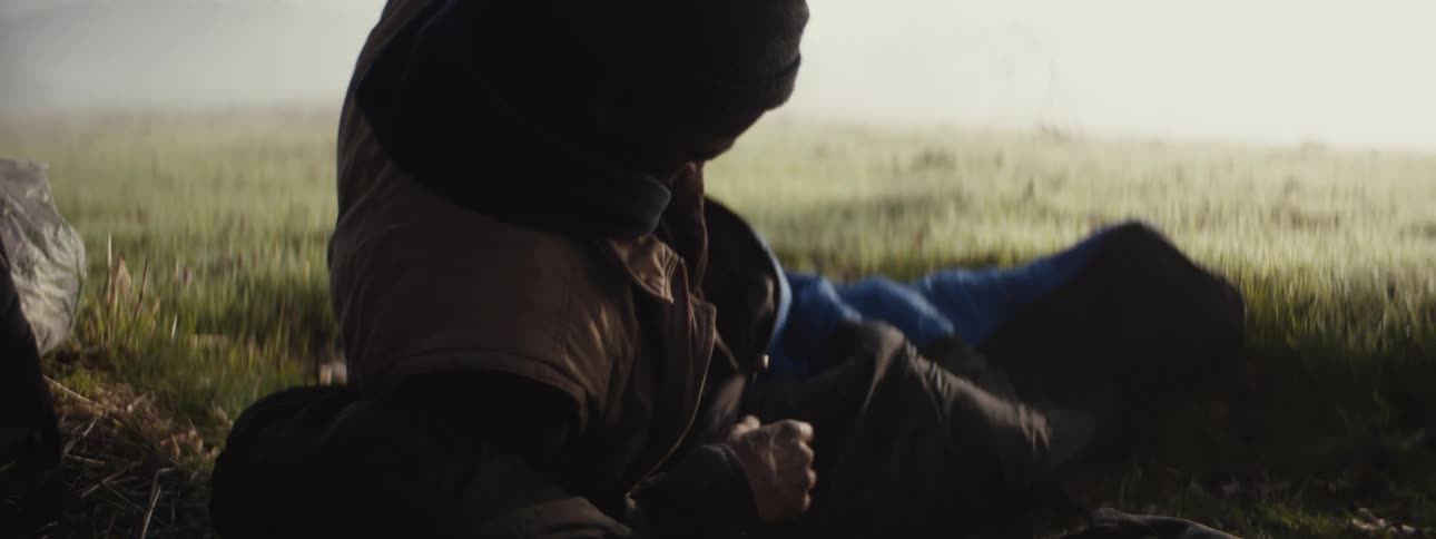 A man sleeping on a sleeping bag in a mist covered grassy field wakes up in the morning