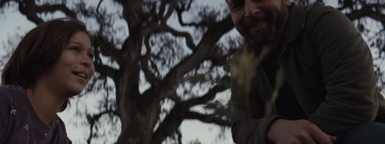 A young girl with short hair and a gentle smile alongside a bearded man in a dark jacket both outdoors near a large twisted tree with a soft moody atmosphere