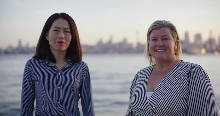 Two women wearing striped shirts are standing in front of a waterfront with a city skyline in the background