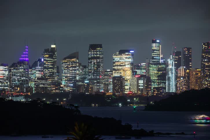 A cityscape skyline glows with lights at night reflected in the dark water with blurred boat lights in the foreground