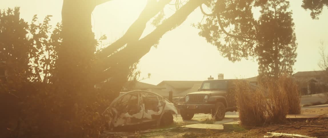 A damaged lightcolored car is parked in a sunny outdoor setting next to a darkcolored jeep with trees and houses visible in the background