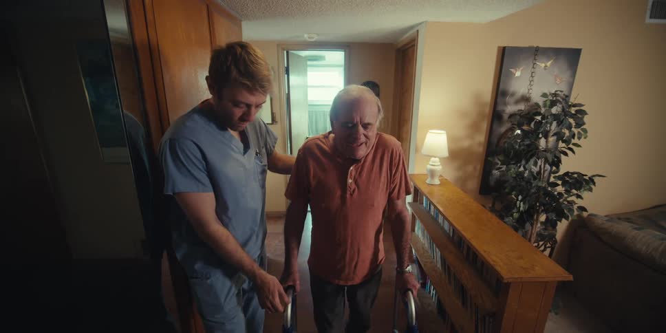 Nurse assisting an elderly man as he walks with the aid of a walker down the hallway of a home