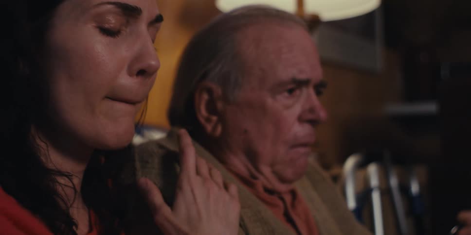 A woman with her eyes closed caressing an elderly man who is sitting next to her and is worried