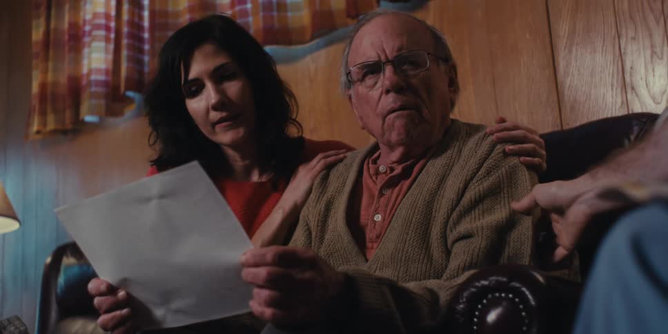 A woman consoles an elderly man who is reading a document with a concerned expression at home