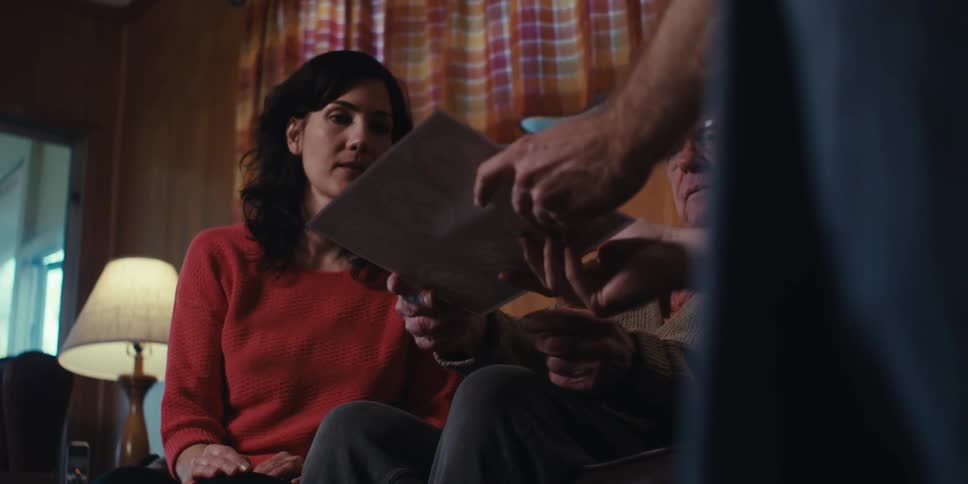 Document being handed to an elderly man who is sitting on the couch next to a woman