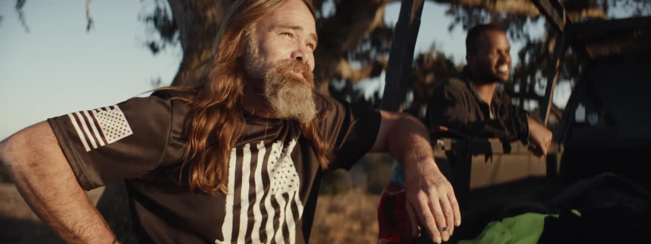 A longhaired and bearded man is leaning against an open car door and talking to a man behind him