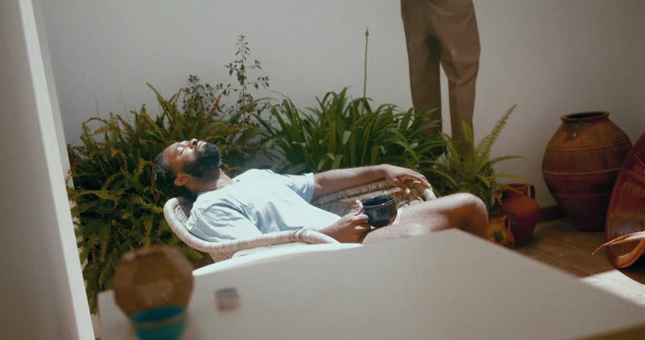 A bearded man wearing a light blue t shirt and orange shorts reclines in a chair near the plants holding a cup of coffee