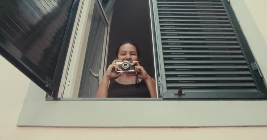 A woman is standing at an open window smiling while holding a camera and taking a photograph