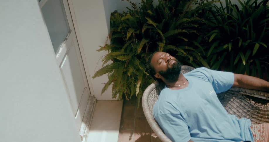 Bearded man reclines in a chair near the houseplants drinking a cup of coffee