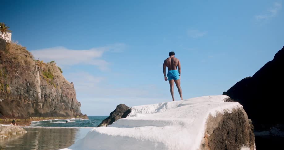 A person in blue shorts is standing on a white stone pathway that leads to the ocean with rocky cliffs and clear blue sky in the background