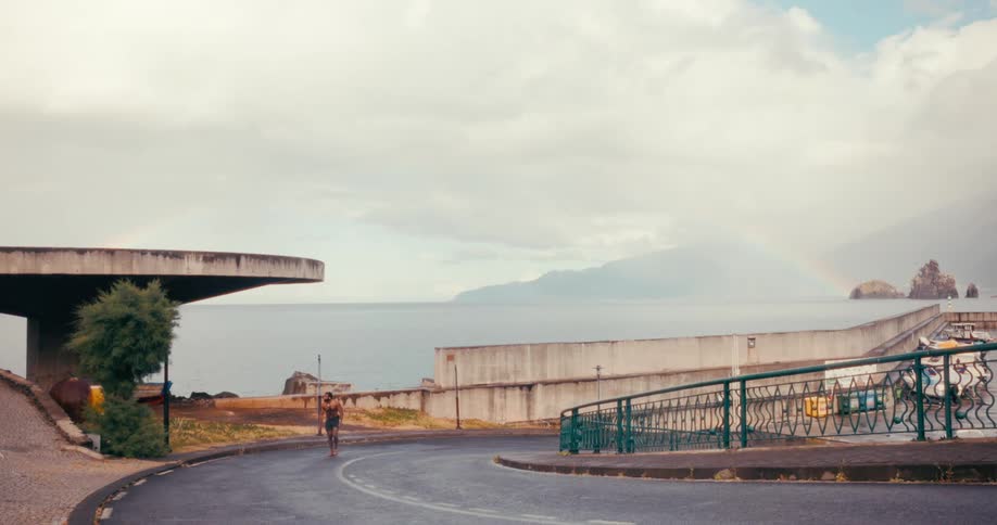 A shirtless muscular man is climbing a curved road near a coastal area with concrete structures near the ocean under a cloudy sky