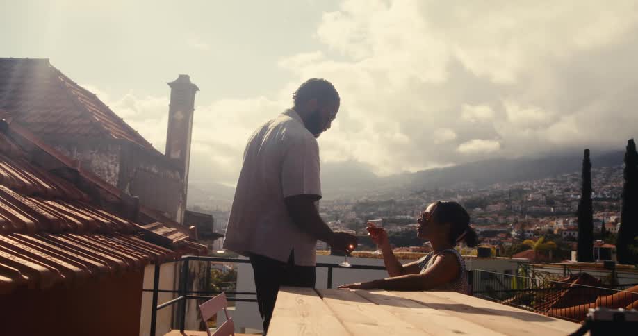 A couple clinks their wine glasses together at a wooden table on a rooftop terrace overlooking a picturesque mountainous cityscape