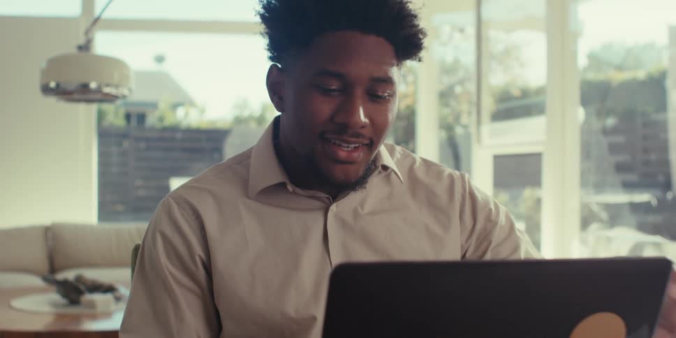A person is sitting at a table in a bright modern room with large windows gesturing while on a video call using a laptop