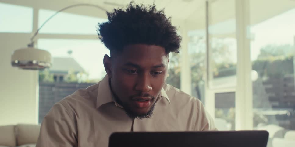 A man in a brown shirt sits at a table in his living room at home using his laptop and becoming excited