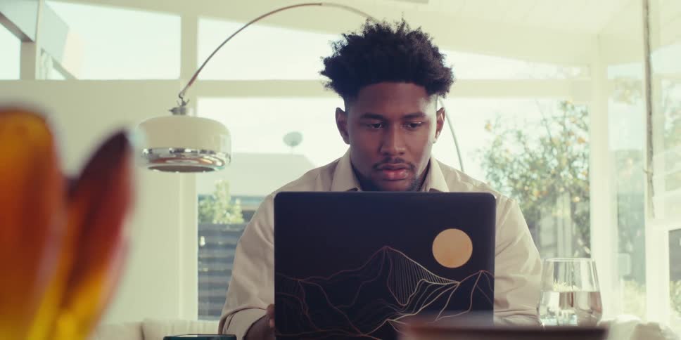 Pensive man wearing a brown shirt sits at table and uses a laptop in the living room