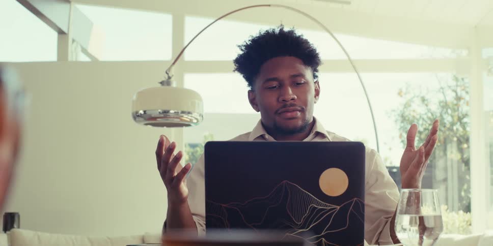 A person is sitting at a table in a bright modern room with large windows gesturing with both hands while on a video call using a laptop
