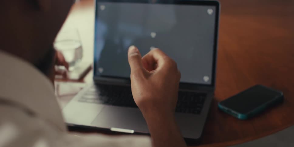 A black man claps his hands while sitting at a wooden table and staring at his laptop screen