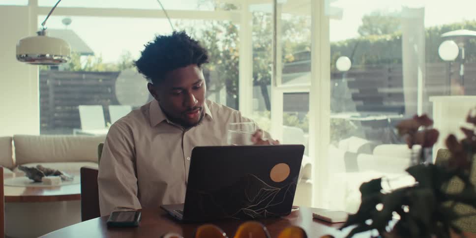 A person is sitting at a table working on a laptop in a bright modern white room with large windows that overlook a patio